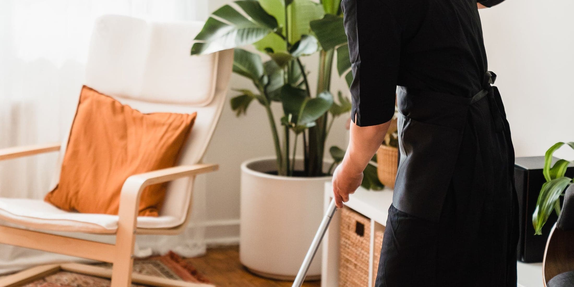 Personne en tablier de travail, vue de dos, passant le balai dans un séjour clair